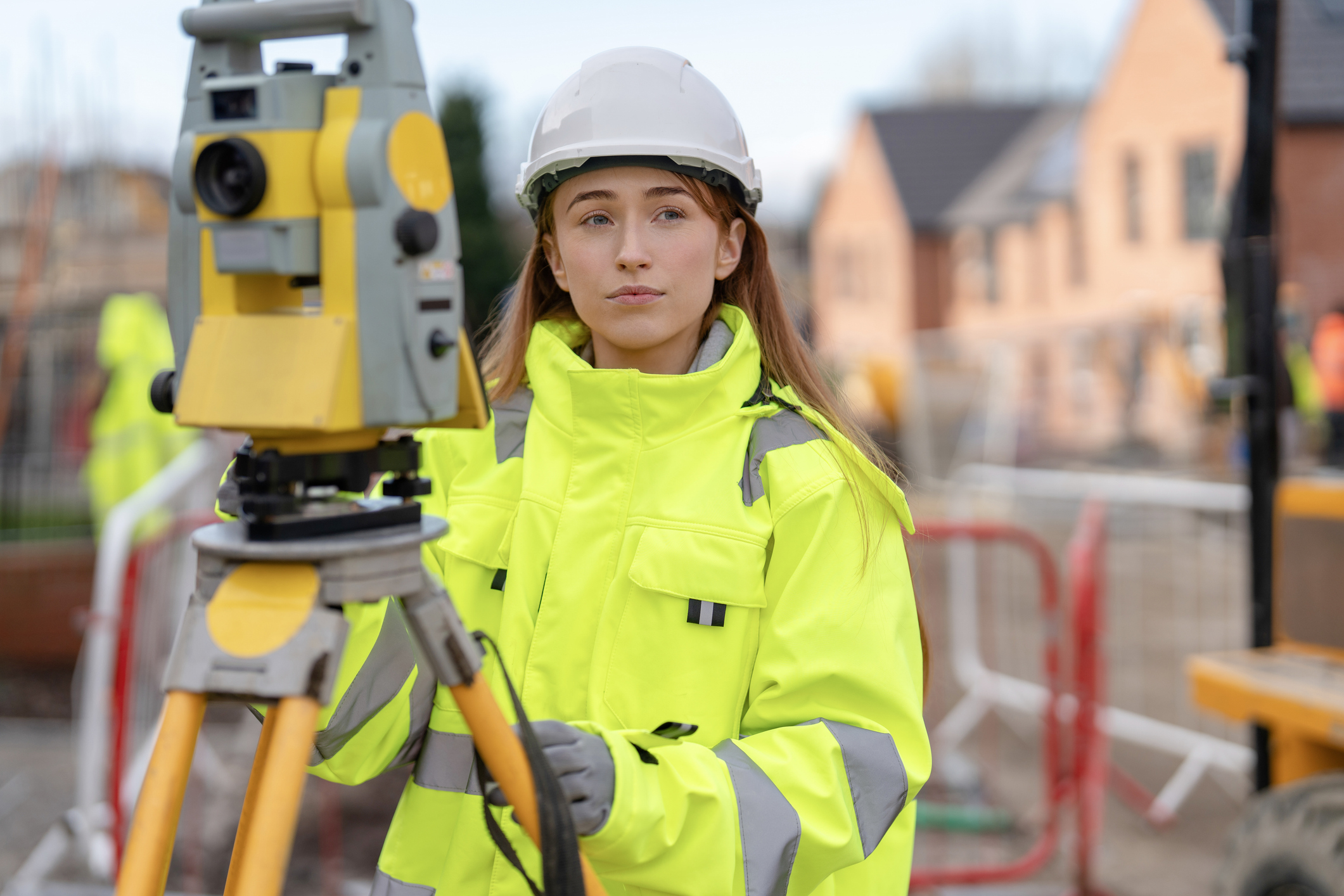 Female Construction surveyor using surveying equipment on building site while wearing bright reflective safety gear